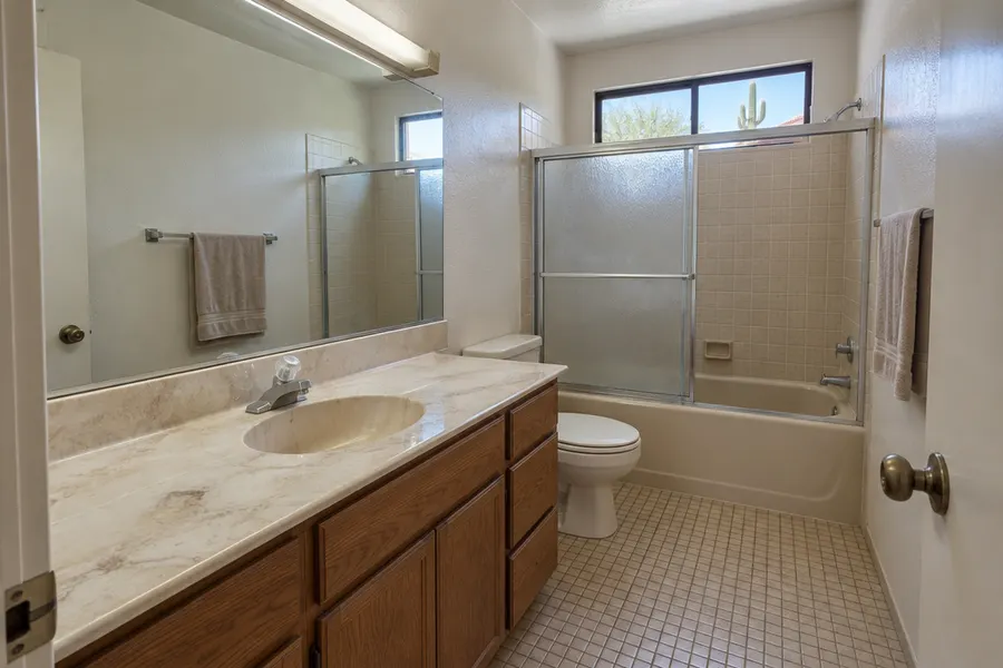 Dated hall bathroom before a remodel: worn cultured-marble vanity, almond bathtub with sliding glass doors, small beige tile and old fluorescent light fixture
